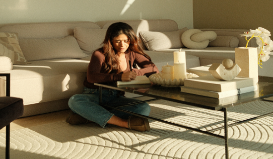 Woman sitting on the floor reading a book in a cozy living room with sunlight filtering through.